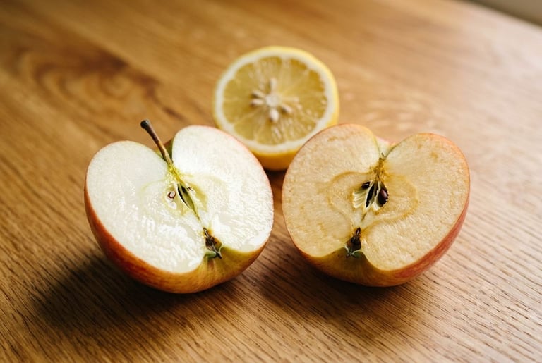 Two halves of the same apple on an oak counter, the left half still bright white and wet from lemon juice and the right half turned an even pale tan across its cut face, with a cut lemon behind them