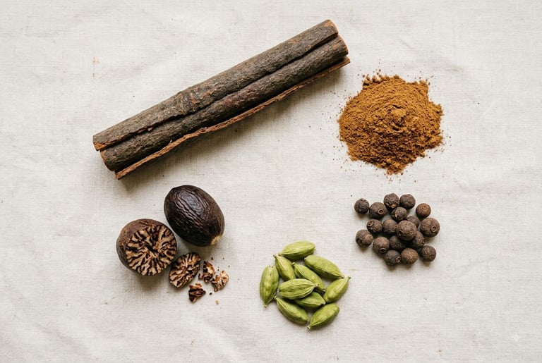 A cassia cinnamon stick and ground cinnamon next to piles of nutmeg, allspice, and cardamom