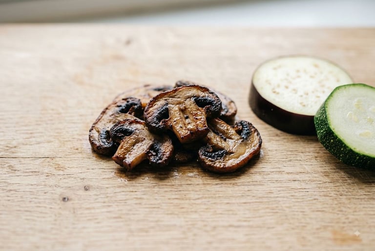 A pile of browned sautéed mushroom slices next to a raw slice of eggplant and a raw slice of zucchini