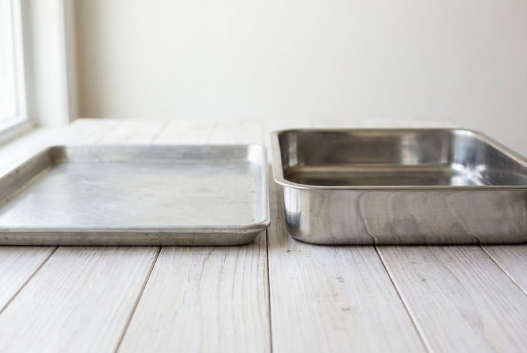 A low-rimmed aluminium sheet pan beside a stainless roasting pan with tall straight walls, on a white-washed wooden counter